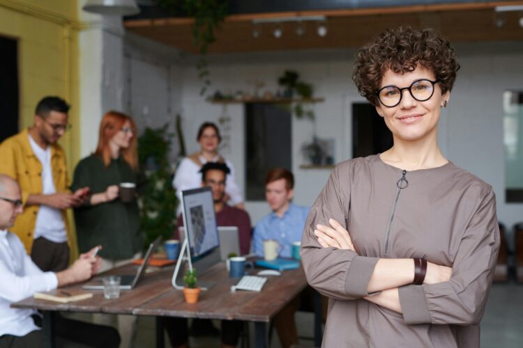 زن با عینک و موهای فرفری که در یک محیط کاری مدرن در مقابل همکاران ایستاده است.